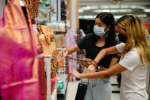 Two women wearing masks shop for traditional jewelry at an indoor market, symbolizing the new normal.